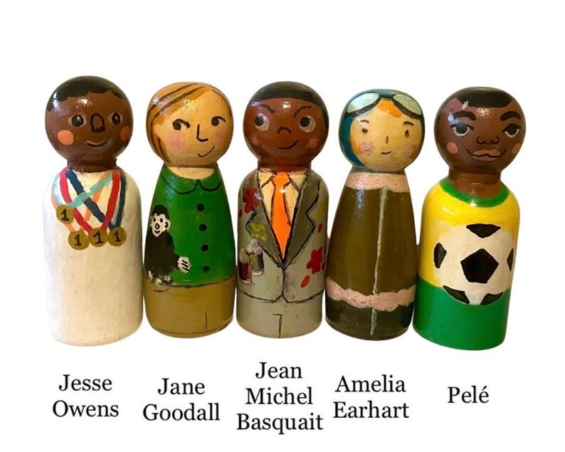 May include: Five hand-painted wooden peg dolls depicting Jesse Owens, Jane Goodall, Jean-Michel Basquiat, Amelia Earhart, and Pel&eacute;. Each doll is uniquely designed with detailed clothing and facial features. The names of each figure are written below them.