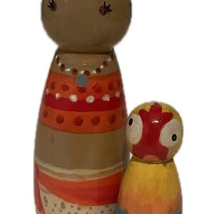 May include: Two hand-painted wooden peg dolls. The larger doll has brown skin, black hair, and is wearing an orange and red dress. The smaller doll has a yellow, orange, and blue body with a red face. Decorative items.