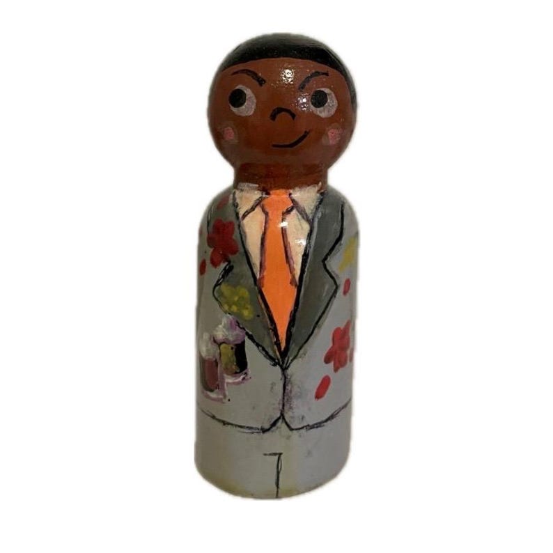 May include: A hand-painted wooden peg doll depicting a person in a gray suit with floral accents and an orange tie. The doll has a dark complexion, black hair, and a smiling expression. The suit has a painted design of beer mugs.