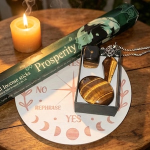 May include: A round wooden board with moon phases and the words NO, REPHRASE, and YES, featuring a box containing a tiger's eye pendulum, a polished tiger's eye stone, and a small black cube, alongside a box of Prosperity incense sticks and a candle.