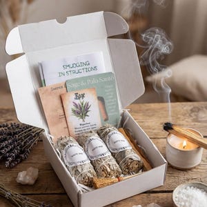 Könnte beinhalten: Eine offene weiße Schachtel mit drei Bündeln weißem Salbei, Anweisungen und Karten. Die Salbeibündel haben weiße Etiketten. Rauch steigt von einem angezündeten Palo Santo-Stab und einer Kerze auf.