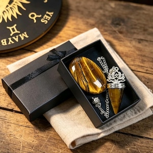 May include: A black gift box containing a polished oval tiger's eye stone and a pointed tiger's eye pendant on a silver-toned chain. The pendant features a silver-toned lotus flower topper. The box is open, revealing the items inside.