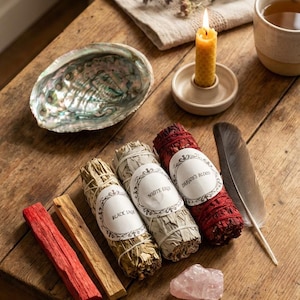 May include: Abalone shell, sage bundles labelled "Black Sage", "White Sage", and "Dragon's Blood", a feather, rose quartz crystal, and a lit beeswax candle on a wooden table. A cup of tea and dried herbs are also present.