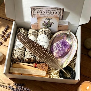 May include: A white gift box containing three bundles of white sage, a brown and white striped feather, a purple amethyst crystal, an iridescent abalone shell, a small broom, a selenite wand, and informational cards about palo santo and sage.