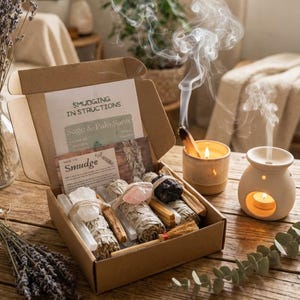May include: A cardboard box containing sage bundles, palo santo sticks, a rose quartz crystal, and a dark stone. Included are instruction cards labelled Smudging Instructions and How to Smudge. Nearby are a lit candle and a ceramic oil burner.