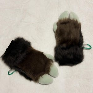 May include: Two faux fur paw gloves. The gloves are brown and black with light green toes. Each glove has a black cuff and a green metal clip.
