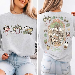 May include: A light gray sweatshirt featuring a colorful graphic of Mickey Mouse, Minnie Mouse, and other Disney characters. The front of the sweatshirt has Mickey Mouse ears, a headband, and coffee cups. The back has a larger graphic of the characters.