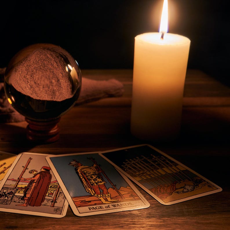 May include: A lit white candle and a crystal ball sit on a wooden table next to tarot cards. The cards feature illustrations and the words "Page of Wands". The scene is dimly lit, creating a mystical atmosphere.
