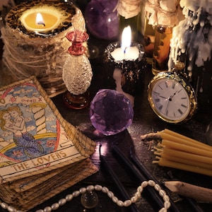 May include: A still life arrangement featuring lit candles, a pocket watch, tarot cards, and crystals. The tarot card reads "The Lovers" and depicts two mermaids. A small glass bottle and a strand of pearls are also present.