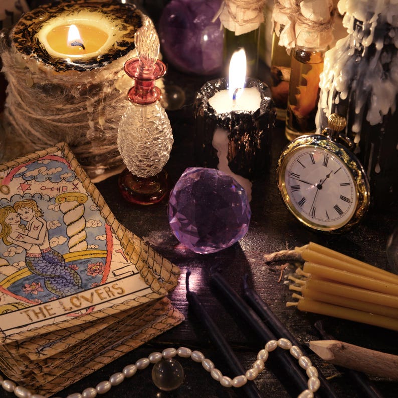 May include: A still life arrangement featuring lit candles, a pocket watch, tarot cards, and crystals. The tarot card reads "The Lovers" and depicts two mermaids. A small glass bottle and a strand of pearls are also present.