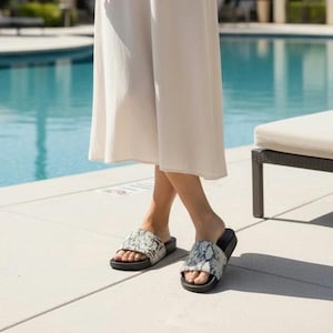 Op de afbeelding: Een persoon draagt een witte broek en instappers met slangenprint en zwarte zolen. De sandalen hebben een brede band over de bovenkant van de voet. De achtergrond omvat een zwembad en een ligstoel.