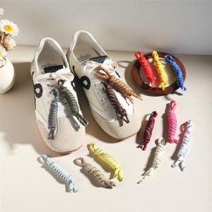 May include: White sneakers with black detailing and beige toe caps are shown with a variety of colourful shoelaces. The shoelaces are in shades of red, yellow, blue, and pink. Some shoelaces are on a wooden plate.