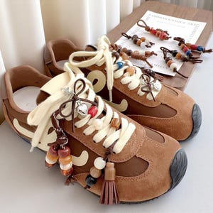 May include: Brown and beige sneakers with white laces and decorative beaded charms. The shoes have a retro design with a suede-like texture and a black toe cap. The laces are adorned with heart-shaped charms and colorful beads.