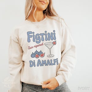 May include: Ivory-colored sweatshirt with the text "Figtini Di Amalfi" in blue and red. The design includes illustrations of figs and a cocktail glass with a fig garnish. The sweatshirt is a casual style.