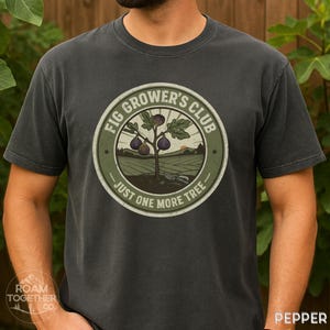 May include: Dark grey t-shirt with a circular graphic featuring a fig tree and the text "FIG GROWER'S CLUB - JUST ONE MORE TREE." The design includes a fig tree with fruit, a field, and a sunset. The shirt has short sleeves and a crew neck.