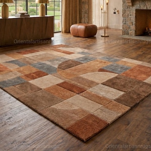 Op de afbeelding: Een groot, rechthoekig vloerkleed met een geometrisch patroon in bruin-, beige- en grijstinten. Het vloerkleed heeft een mix van vierkante en gebogen vormen, wat een modern ontwerp creëert. Het vloerkleed ligt op een houten vloer in een woonkamer.