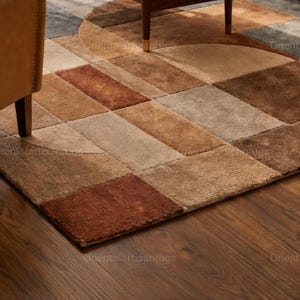 Op de afbeelding: Een geometrisch patroon tapijt in bruin, beige en roest tinten. Het tapijt heeft een patchwork ontwerp met rechthoekige en halfronde vormen. Het tapijt ligt op een houten vloer, met een deel van een stoel zichtbaar.