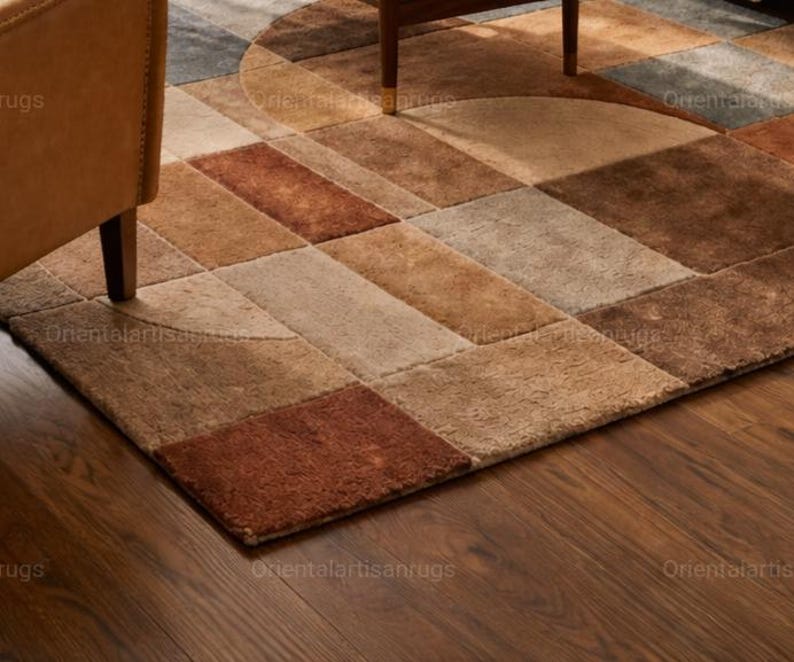 Op de afbeelding: Een geometrisch patroon tapijt in bruin, beige en roest tinten. Het tapijt heeft een patchwork ontwerp met rechthoekige en halfronde vormen. Het tapijt ligt op een houten vloer, met een deel van een stoel zichtbaar.