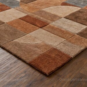Op de afbeelding: Een geometrisch vloerkleed met een patchwork-ontwerp in bruin-, beige- en grijstinten. Het vloerkleed heeft een verscheidenheid aan rechthoekige en halfronde vormen, waardoor een gestructureerd oppervlak ontstaat. Het vloerkleed ligt op een donkere houten vloer.