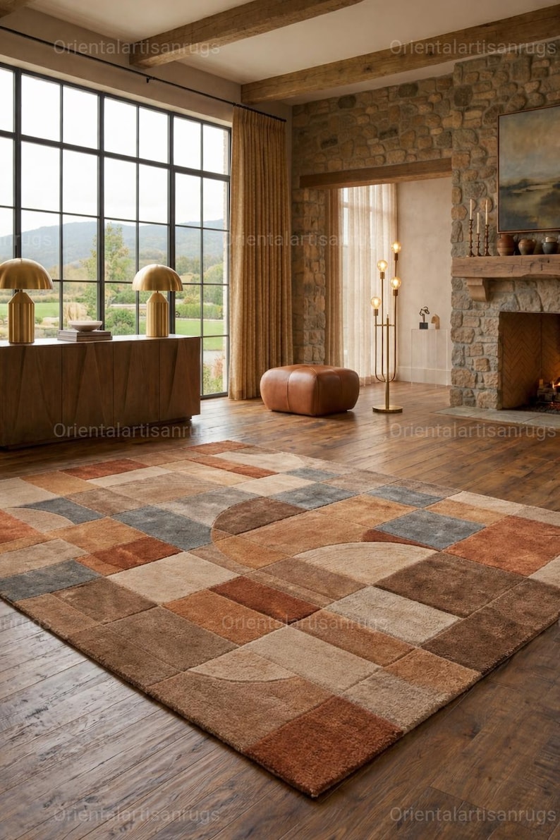 Op de afbeelding: Een groot, rechthoekig vloerkleed met een geometrisch patroon in bruin-, beige- en grijstinten. Het vloerkleed heeft een mix van vierkante en gebogen vormen, wat een modern ontwerp cre&euml;ert. Het vloerkleed ligt op een houten vloer in een woonkamer.