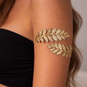 May include: Gold-colored arm cuff featuring a leaf design. The cuff wraps around the upper arm, with two separate leaf-shaped sections connected by a small band. The leaves have detailed veining, creating a realistic appearance. The jewelry is worn on a person's arm.