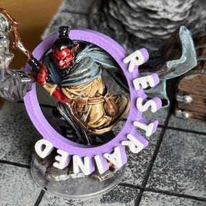 Pu&ograve; includere: Una miniatura di un personaggio demoniaco dalla pelle rossa in un anello viola con la scritta "REST TRAINED". La figura tiene in mano un bastone. Un forziere di legno &egrave; sullo sfondo. La scena &egrave; ambientata su una superficie grigia.