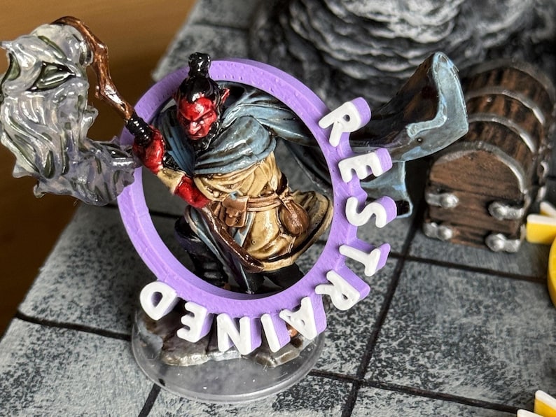 Pu&ograve; includere: Una miniatura di un personaggio demoniaco dalla pelle rossa in un anello viola con la scritta "REST TRAINED". La figura tiene in mano un bastone. Un forziere di legno &egrave; sullo sfondo. La scena &egrave; ambientata su una superficie grigia.