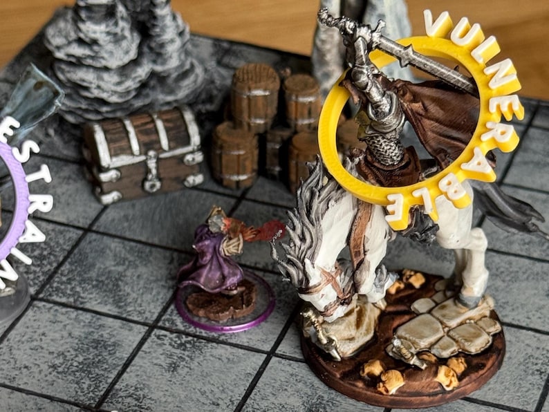 Pu&ograve; includere: Una scena di gioco di ruolo da tavolo con miniature. Un cavaliere a cavallo con una spada affronta un mago che lancia un incantesimo. La parola "VULNERABLE" &egrave; in un anello giallo. Altri elementi includono un forziere e barili.