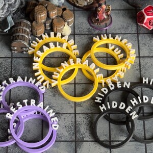 Pu&ograve; includere: Una collezione di accessori per giochi stampati in 3D, tra cui anelli gialli "VULNERABLE", viola "RESTRAINED" e neri "HIDDEN". Dadi e barili in miniatura sono visibili, suggerendo un tema di gioco da tavolo.