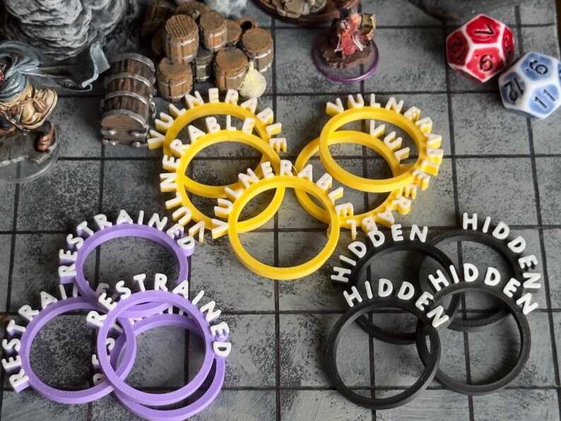 Pu&ograve; includere: Una collezione di accessori per giochi stampati in 3D, tra cui anelli gialli "VULNERABLE", viola "RESTRAINED" e neri "HIDDEN". Dadi e barili in miniatura sono visibili, suggerendo un tema di gioco da tavolo.