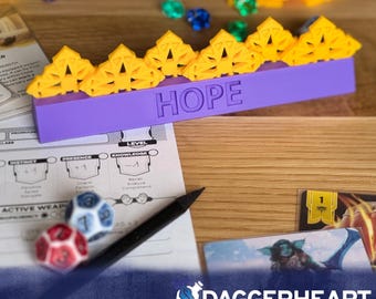 Può includere: Un vassoio per dadi viola con la parola "HOPE" incisa sul lato. Il vassoio contiene porta dadi a forma di corona gialla. Sono visibili anche dadi, carte da gioco e una matita, insieme al testo "DAGGERHEART COMPATIBLE".
