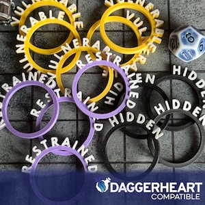 May include: A collection of colorful game tokens with the words "Vulnerable", "Restrained", and "Hidden" printed on them. The tokens are yellow, purple, and black. A blue and white die is also present, along with the text "Daggerheart Compatible".