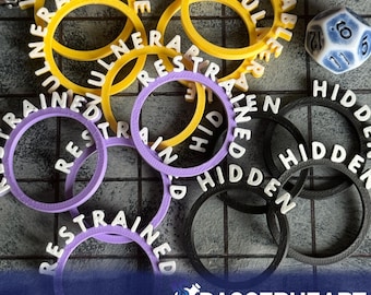 Può includere: Una collezione di gettoni di gioco colorati con le parole "Vulnerable", "Restrained" e "Hidden" stampate sopra. I gettoni sono gialli, viola e neri. È presente anche un dado blu e bianco, insieme al testo "Daggerheart Compatible".