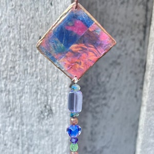 May include: A diamond-shaped hanging ornament with a copper-toned frame containing a multi-colored abstract resin insert in shades of blue, pink, and orange. A vertical strand of assorted glass beads in purple, blue, green, and red hangs below.