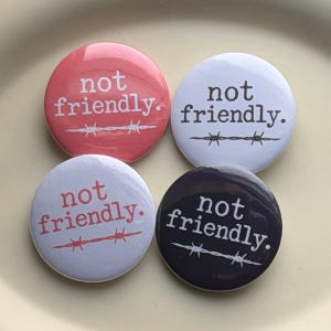 May include: Four circular pinback buttons featuring the text "not friendly." in a serif font above a graphic of barbed wire. The buttons come in four distinct colors: a salmon pink, a white, a light gray, and a dark charcoal gray.