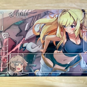 Może przedstawiać: Prostokątna mata do gry w karty Gundam z grafiką w stylu anime przedstawiającą blondwłosą kobietę i mecha. Mata ma wyznaczone obszary do gry, oznaczone jako "BASE", "SHIELDS", "DECK", "TRASH" i "RESOURCE AREA".