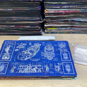 Pode incluir: Tapete de rato azul com um design de planta mostrando um veículo em branco. Uma pequena bolsa branca com cordão e uma caneta também estão presentes. Pilhas de tapetes de rato semelhantes estão no fundo.
