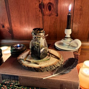 May include: A clear glass bottle with a cork stopper sealed in black wax, containing dark liquid and herbs, sits on a circular wood slice with small twigs. Nearby are a black candle in a white holder, a black feather, and a brown book.
