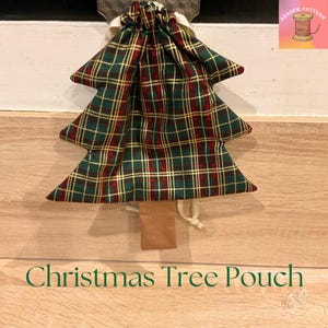 Peut inclure: Une pochette en forme de sapin de Noël en tissu à carreaux vert, rouge et or. La pochette a une fermeture à cordon et une petite étiquette marron à la base. Le texte "Christmas Tree Pouch" est affiché en vert.