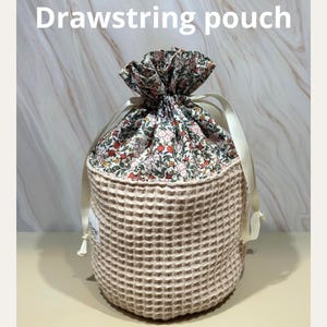 Peut inclure: Un sac à cordon beige avec une texture gaufrée. Le haut est à motifs floraux avec des fleurs roses et rouges. Des rubans crème sont noués en haut. Le texte "Drawstring pouch" est en haut.