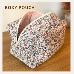Op de afbeelding: Een boxy etui met een bloemenpatroon in roze, rood en groen op een witte achtergrond. De etui heeft een ritssluiting en een roze binnenkant. De tekst "BOXY POUCH" staat bovenaan de afbeelding.