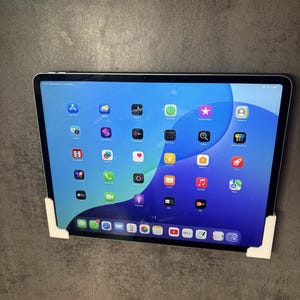 Puede incluir: Una tableta con una pantalla azul y blanca que muestra iconos de aplicaciones, sujeta por soportes de esquina blancos. La pantalla muestra una variedad de iconos de aplicaciones coloridos, incluyendo aplicaciones de redes sociales y entretenimiento. La tableta está montada en una pared gris.