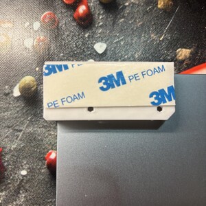 Pu&ograve; includere: Una striscia adesiva di plastica bianca con la scritta "3M PE FOAM" e il logo 3M in blu. La striscia &egrave; attaccata a una superficie metallica grigia. Lo sfondo presenta una superficie scura e strutturata con cibo sparso.
