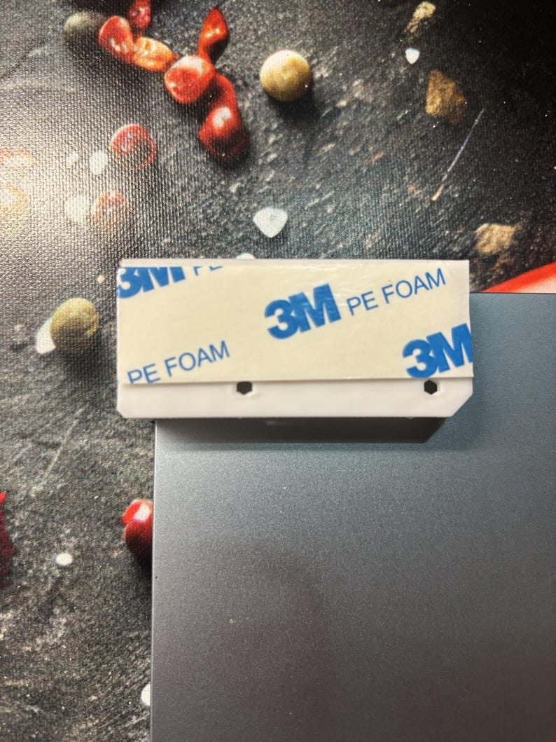 Pu&ograve; includere: Una striscia adesiva di plastica bianca con la scritta "3M PE FOAM" e il logo 3M in blu. La striscia &egrave; attaccata a una superficie metallica grigia. Lo sfondo presenta una superficie scura e strutturata con cibo sparso.