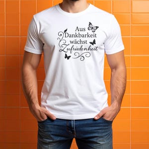 May include: White t-shirt with short sleeves featuring the German phrase "Aus Dankbarkeit w&auml;chst Zufriedenheit" in black script, accompanied by butterfly illustrations. The shirt is worn with blue jeans.