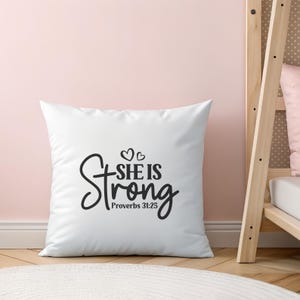 Peut inclure: Coussin carr&eacute; blanc avec l'inscription "She is Strong" en &eacute;criture noire, avec deux petits c&oelig;urs au-dessus du mot "is" et le texte "Proverbes 31:25" en dessous. Fond rose clair.
