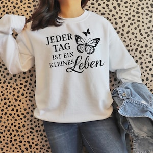 May include: White sweatshirt with long sleeves featuring a black butterfly graphic and the German text "Jeder Tag ist ein kleines Leben." The sweatshirt is paired with blue jeans and a denim jacket.