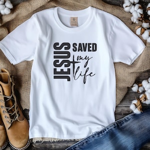 Peut inclure: T-shirt blanc avec l'inscription noire "JESUS SAVED my life". Le mot "JESUS" est empil&eacute; verticalement &agrave; gauche, avec une croix au milieu. Les mots "SAVED my life" sont &agrave; droite. Le t-shirt est pr&eacute;sent&eacute; sur une surface en bois.
