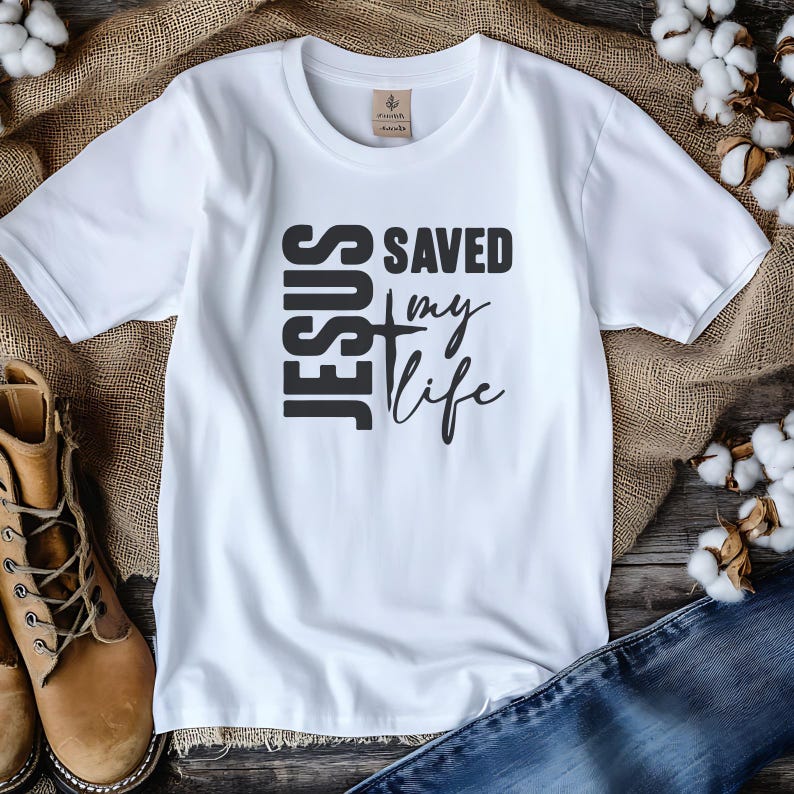 Peut inclure: T-shirt blanc avec l'inscription noire "JESUS SAVED my life". Le mot "JESUS" est empil&eacute; verticalement &agrave; gauche, avec une croix au milieu. Les mots "SAVED my life" sont &agrave; droite. Le t-shirt est pr&eacute;sent&eacute; sur une surface en bois.