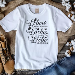 May include: White t-shirt with a crew neck featuring a black script design with butterflies and the words "Leben jeden Moment Lache jeden Tag Liebe unendlich". The shirt is made of a soft material.
